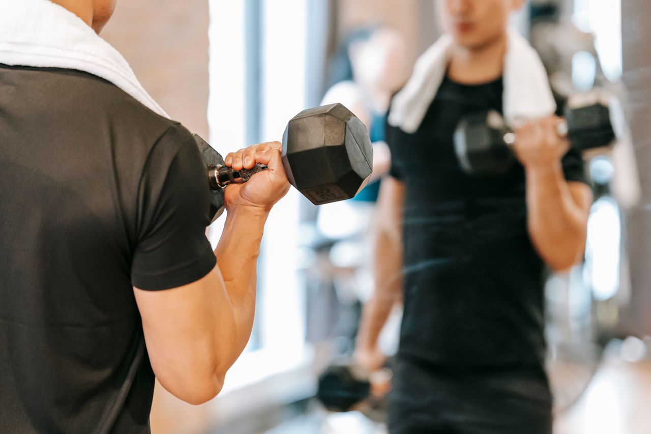 Unrecognizable male athlete with towel pumping biceps with dumbbells while standing near mirror during fitness training in gym on blurred background